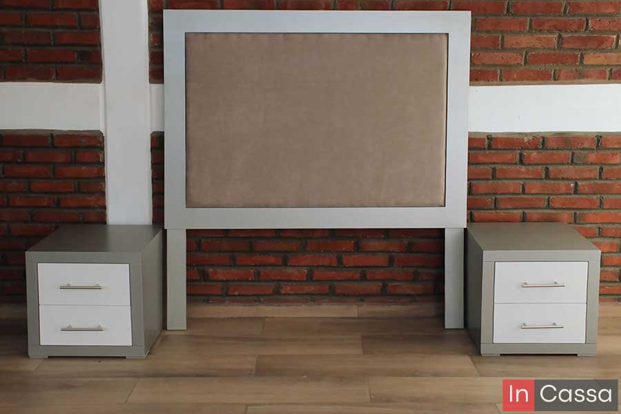 En esta toma frontal se aprecia la cabecera tapizada en suede camel con marco blanco, acompañada por dos burós color silver con cajones blancos y jaladeras metálicas. El fondo de ladrillo y piso tipo madera acentúan su estilo moderno y equilibrado.
