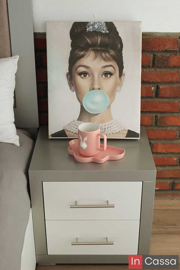 La foto presenta un buró en color silver con dos cajones blancos, jaladeras metálicas y diseño moderno. Encima se exhibe una taza rosa y un cuadro decorativo que acentúan el estilo juvenil y elegante del espacio. El muro de ladrillo agrega textura al entorno.