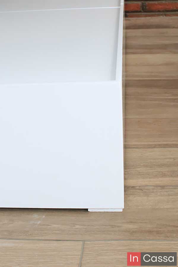 Se observa el diseño minimalista del frente de la base, con líneas rectas y bordes definidos. El acabado blanco brillante resalta sobre el piso tipo madera, y la base se eleva ligeramente gracias a discretos niveladores.