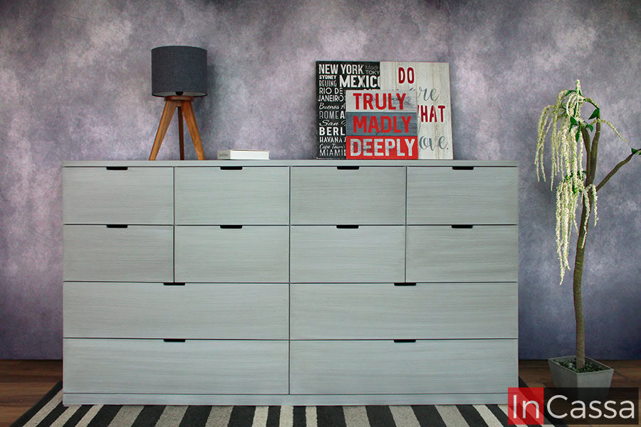 Cómoda - Cajonera tamaño jumbo en color gris cenizo claro con 12 cajones de diferentes tamaños. El mueble es presentado con algunos artículos decorativos en una habitación con pared gris y piso de madera.