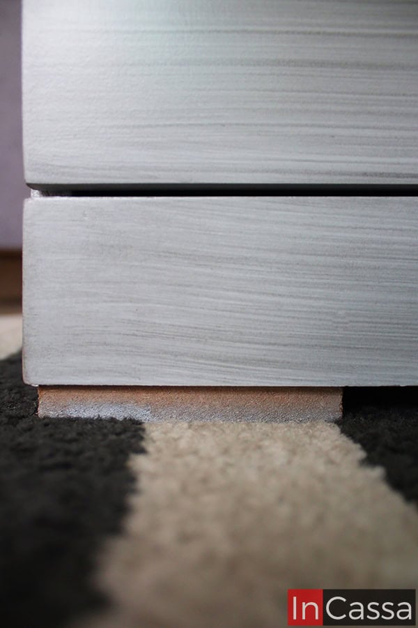 La foto se concentra en la base de la cajonera, destacando su acabado gris cenizo claro así como una de las patas planas de madera que sostienen al mueble.