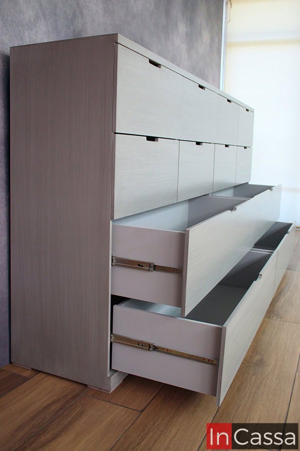 Toma lateral de la cómoda - cajonera con sus cajones inferiores abiertos, en la que se puede apreciar la estructura completa de cada compartimento así como de su sistema de apertura y cierre conformado por rieles metálicos. El interior de cada cajón es de un tono gris similar al de las partes externas del mueble en su totalidad.