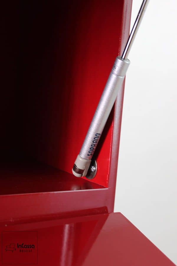 La imagen muestra el sistema de pistón a gas que facilita la apertura suave y segura de las puertas de las alacenas superiores. Su acabado metálico contrasta elegantemente con el interior rojo brillante del mueble, destacando la calidad de los herrajes.