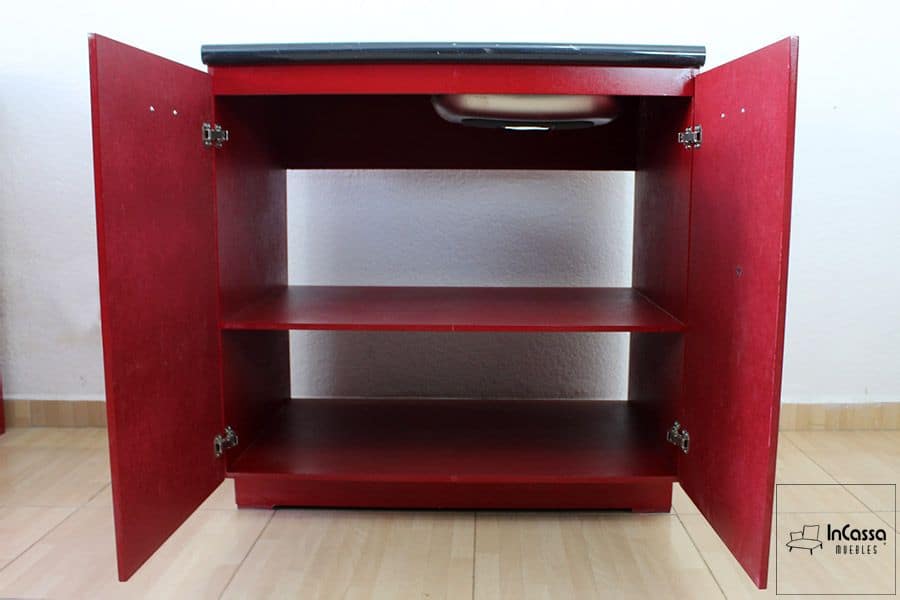 Este gabinete inferior en tono rojo quemado muestra su interior con repisa fija para almacenamiento dividido. Las bisagras metálicas aseguran durabilidad, mientras que la cubierta superior negra sostiene la tarja de acero inoxidable, visible parcialmente desde abajo.