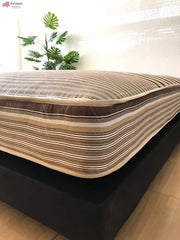 Vemos el colchón hotelero, con un estilo de rayas en el acolchonado, presentado sobre una base tapizada negra en una habitación con pared blanca y piso de madera.