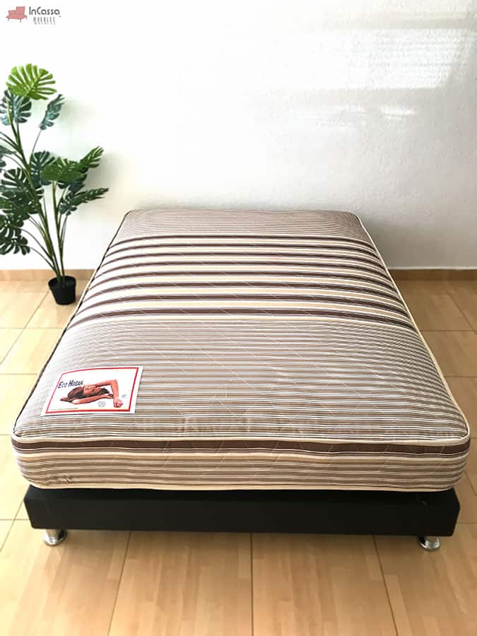 Una imagen completa del acolchonado del colchón en el que vemos un notable estilo de rayas así como la etiqueta de presentación. La presentación se realizo en una estancia con pared blanca y piso de madera, el colchón es acompañado por una base tapizada negra con patas metálicas.