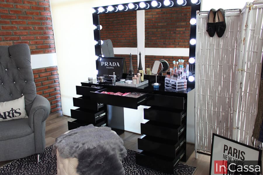 Una foto que nos muestra una toma lateral del tocador, en la cual podemos apreciar mejor los el espejo con sus 18 focos encendidos, los cuales ayudan a resaltar el acabado negro brillante del mueble. Además se nos presenta el tocador con todos sus compartimentos dejándonos ver su estructura completa.