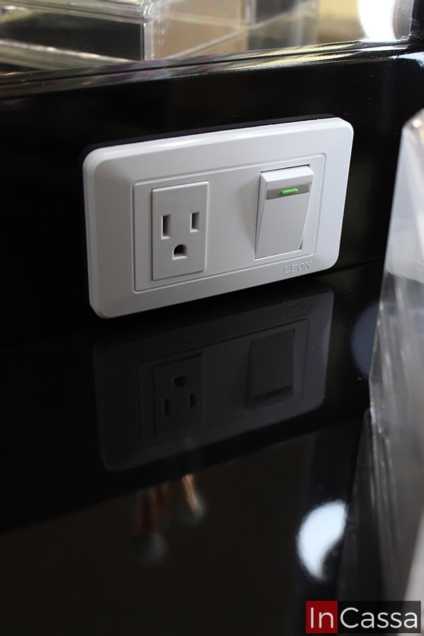Una imagen del contacto eléctrico incorporado al espejo del tocador. El cual cuenta con apagador para encender los 18 focos y enchufe para conectar cualquier dispositivo eléctrico. Por otro lado, la toma también permite observar parte de la cubierta y el marco del espejo, ambos en acabado negro brillante.