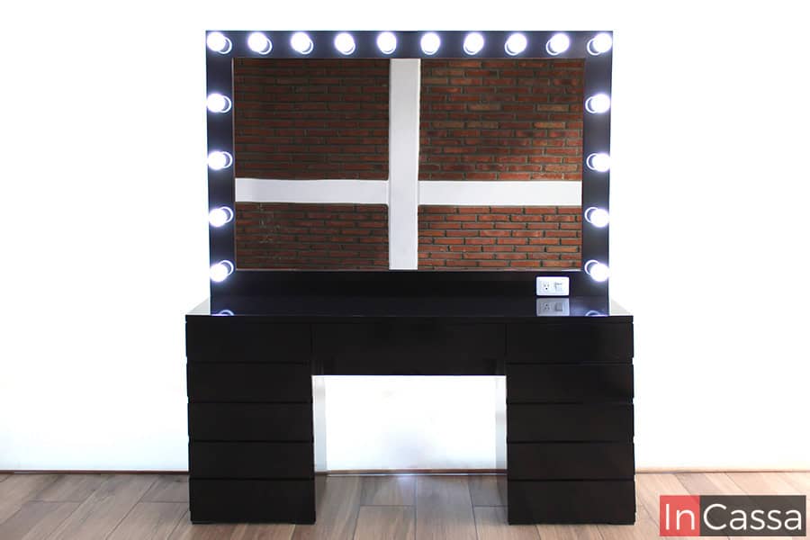 Vemos una foto completa del tocador negro con espejo de 18 focos, los cuales se presentan encendidos resaltando el acabado brillante de la pintura. El mueble se encuentra instalado en una estancia con ventana de pantalla completa y piso de madera.