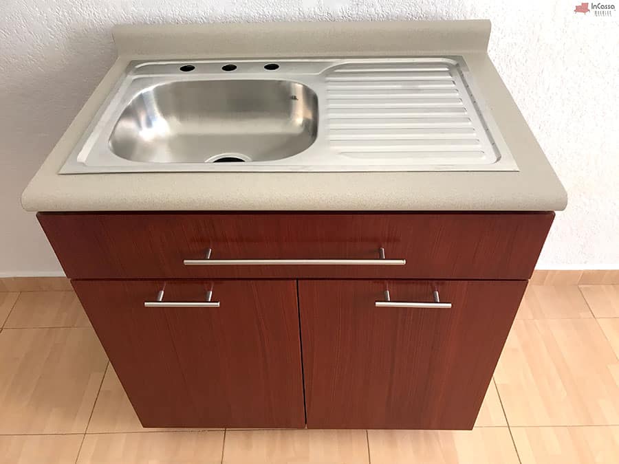 La foto presenta el módulo inferior con tarja integrada. Este módulo de color maple oscuro combina funcionalidad y diseño con una tarja de acero inoxidable y una amplia cubierta de formaica estilo granito de arena para tareas de cocina.