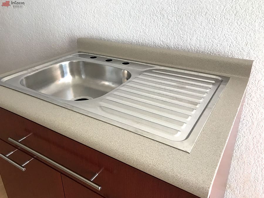 La foto presenta el diseño de la tarja de acero inoxidable. Este elemento incluye un escurridor amplio y funcional, complementado por una cubierta de formaica en tono granito de arena para mayor durabilidad.