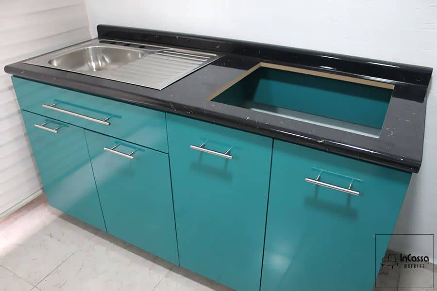 Esta toma lateral del módulo inferior de la cocina, nos proporciona una vista completa del mueble, en el que podemos ver el espacio para parrilla así como la tarja de acero inoxidable instalada en la cubierta de formica estilo mármol negro. Sin mencionar que también se nos muestra las puertas y estructura del mueble presentados en un acabado turquesa brillante.