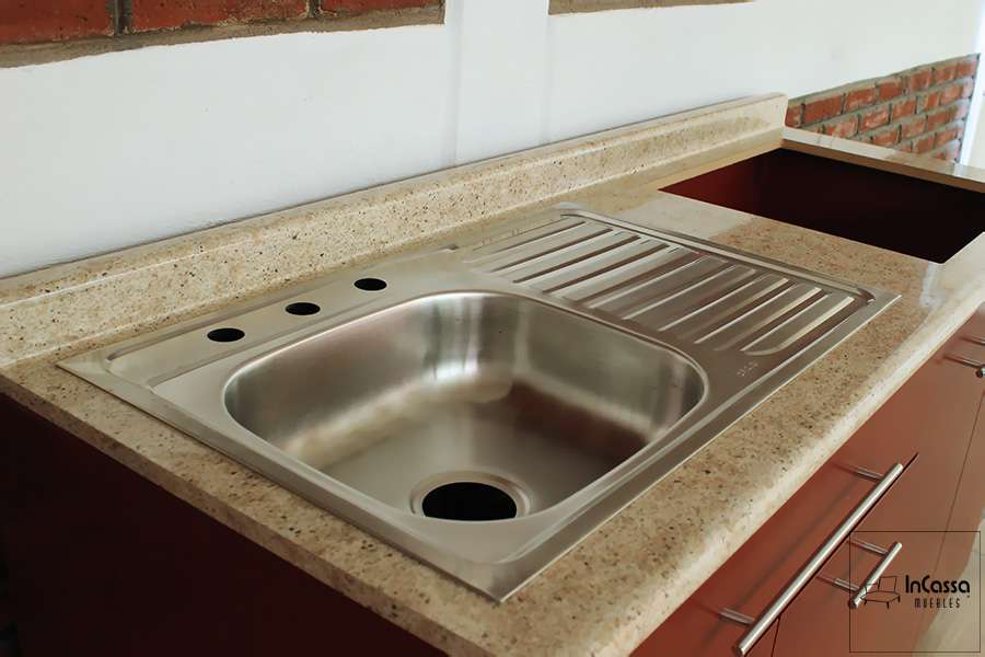 Una toma lateral del módulo inferior de la cocina, en el cuál destaca la tarja de acero inoxidable añadida en la cubierta de formica estilo ojo de pájaro, la tarja cuenta con tina izquierda y escurridor derecho- También se puede ver el hueco para parrilla así como parte de las puertas y estructura de color maple oscuro, además de sus jaladeras metálicas integradas.