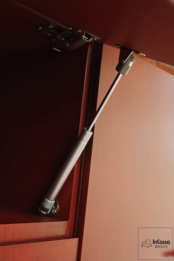 En la imagen vemos el sistema de apertura y cierre instalado en los módulos superiores de la cocina, el cuál consiste en un pistón neumático metálico. Adicionalmente podemos apreciar una pequeña parte del interior del compartimento, así como una vista más detallada de su acabado maple oscuro.