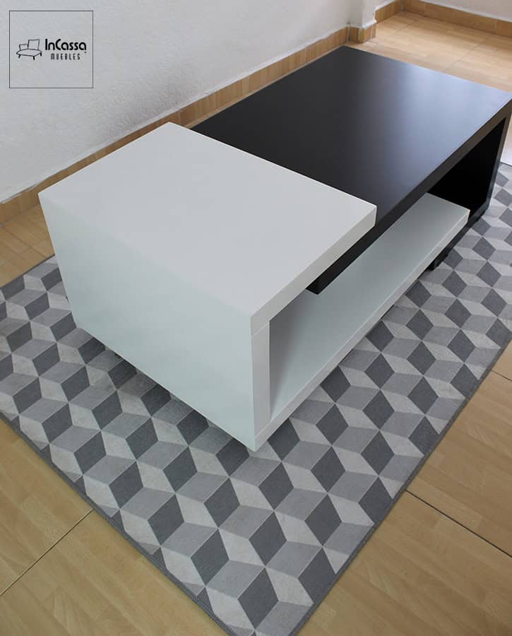 Desde una vista en picada se resalta el diseño en L invertida del módulo blanco, que sobresale sobre el tablero negro. La fotografía también deja ver la amplitud del estante inferior y cómo el mueble se integra con elegancia sobre una alfombra de patrón geométrico.
