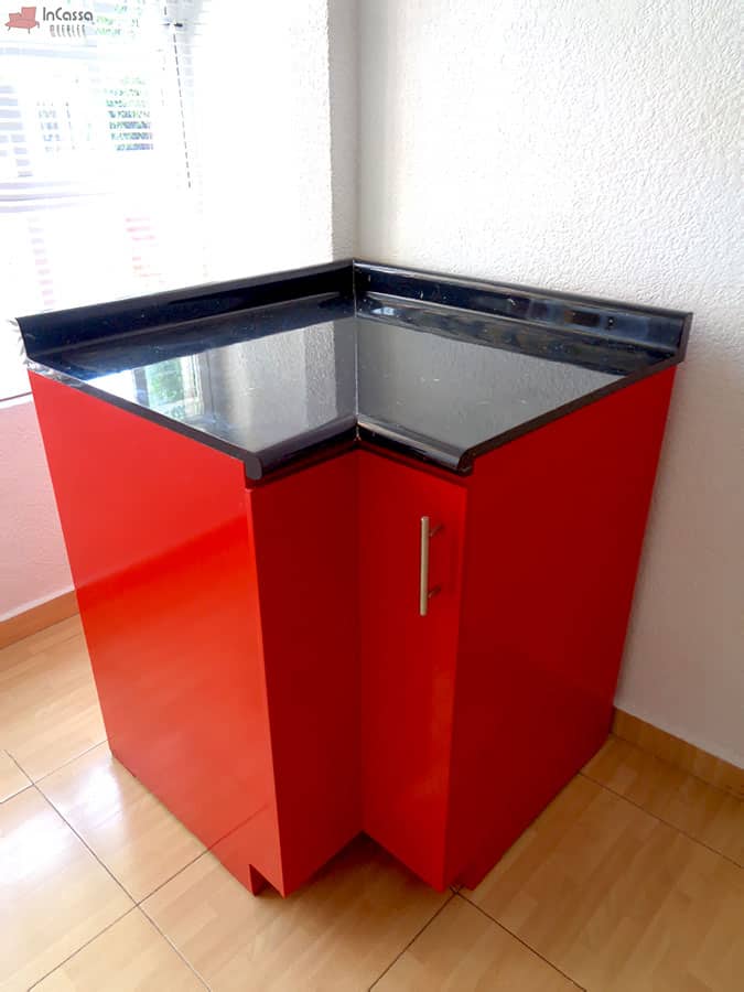 Fotografía en ángulo que muestra el módulo esquinero de cocina Italia con puerta frontal roja de acabado brillante, jaladera metálica vertical y cubierta de formaica tipo mármol negro con salpicadera integrada. La superficie refleja la luz natural que entra por la ventana cercana, resaltando el contraste moderno entre el rojo intenso y el negro pulido.