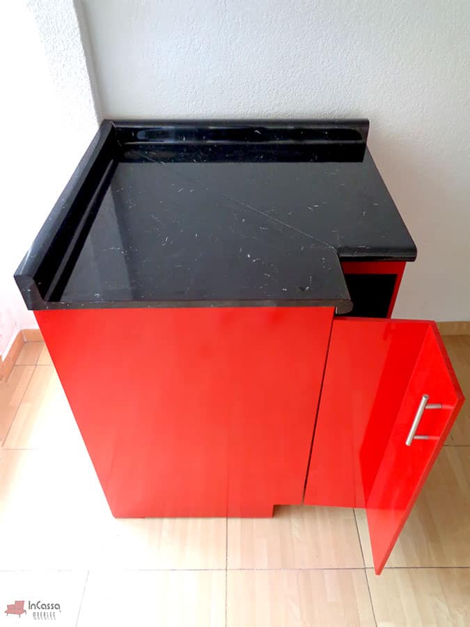 Imagen tomada desde arriba que muestra el módulo esquinero de cocina Italia en color rojo brillante, con puerta lateral abierta y jaladera metálica vertical. La cubierta de formaica estilo mármol negro con salpicadera presenta un diseño en corte diagonal para ajustarse a la esquina, aportando un estilo moderno y funcional.