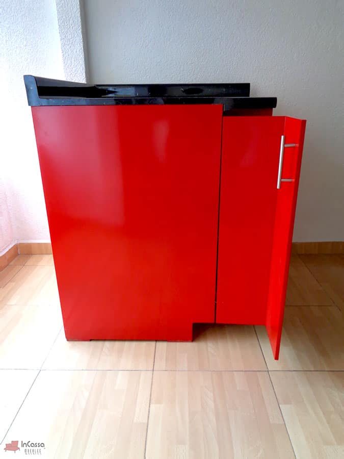 Toma lateral que muestra el módulo esquinero de cocina Italia en acabado rojo brillante, con puerta lateral abierta y jaladera metálica vertical. La cubierta de formaica estilo mármol negro con salpicadera superior contrasta con el diseño moderno del mueble, resaltando su estructura compacta y funcional.