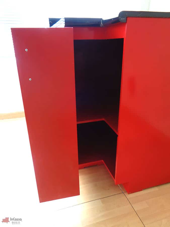 Toma frontal que revela el interior del módulo esquinero Italia en acabado rojo brillante, con puerta abierta mostrando un entrepaño interno en color rojo. La cubierta superior de formaica en estilo mármol negro con salpicadera resalta el contraste de colores, aportando un diseño elegante y funcional para almacenamiento.