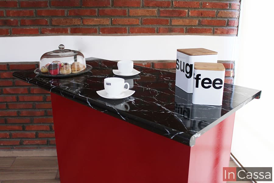 La imagen muestra una toma cercana de la barra de cocina destacando la cubierta de formica estilo mármol negro, misma que es visiblemente más amplia que la base roja ofreciendo un mayor espacio para colocar vajillas y utensilios.