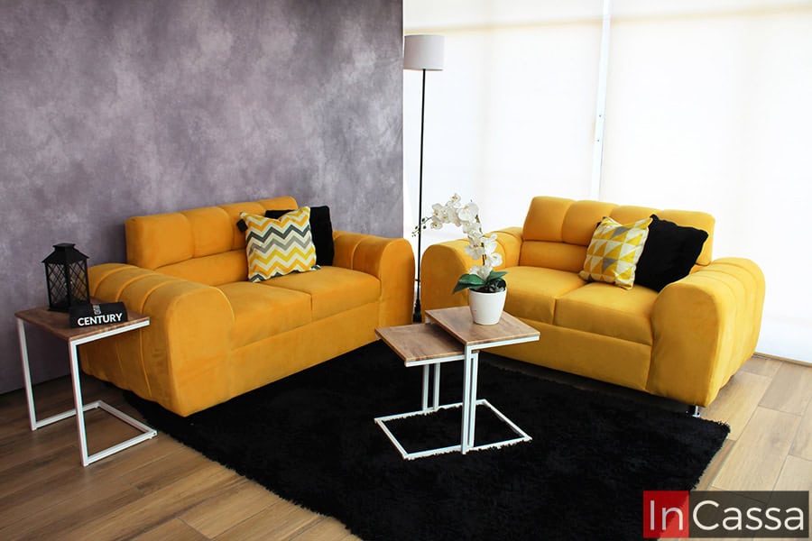 La imagen muestra una sala de estar minimalista con dos love seat en vibrante suede color dorado, decorados con cojines geométricos. Las mesas de centro modernas y la alfombra negra crean un ambiente acogedor y elegante.