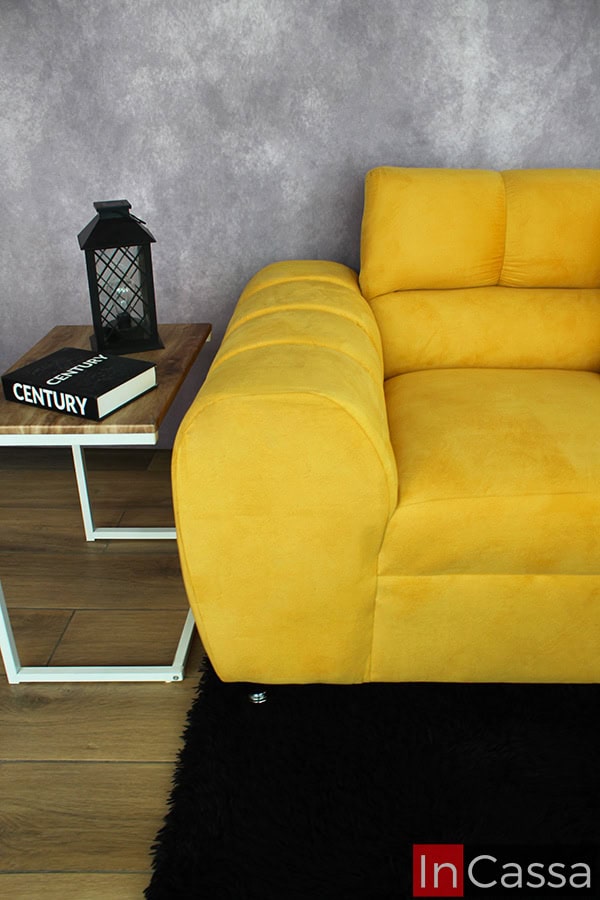 Una toma de un love seat de suede dorado vibrante, acompañado de una mesa lateral moderna con una lámpara decorativa y un libro. La combinación de colores y texturas crea un ambiente contemporáneo y acogedor.