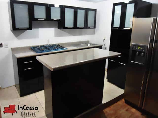 Se presenta una cocina integral en acabado negro brillante con puertas de vidrio esmerilado en la parte superior y barra desayunador central con cubierta de formaica en estilo mármol escalona. A la derecha destaca una alacena alta que brinda gran capacidad de almacenamiento, junto a un refrigerador de doble puerta en acero inoxidable que armoniza con el diseño moderno del espacio.