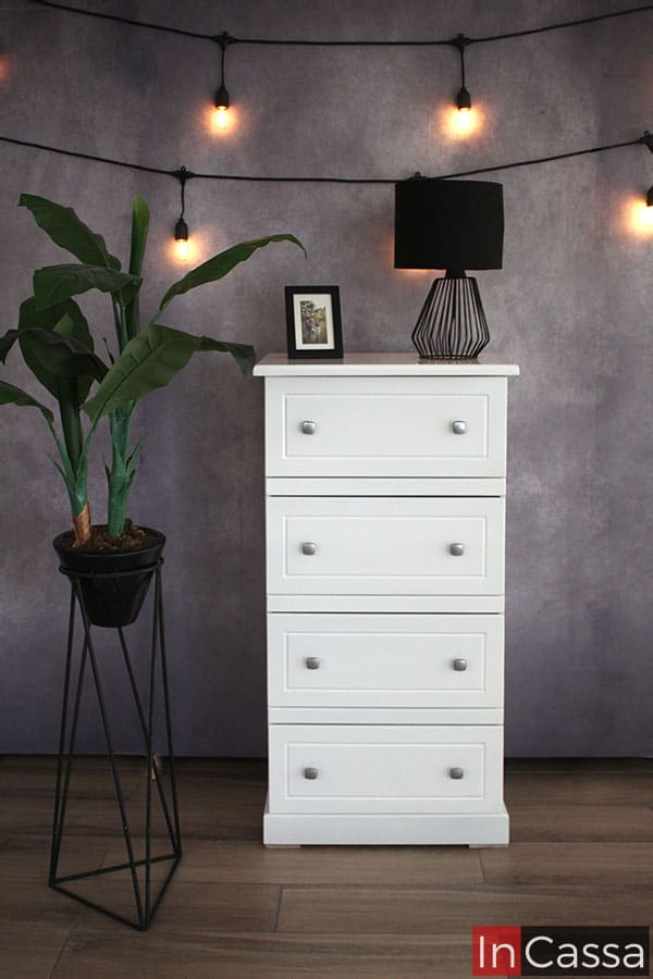 Una toma frontal de la cajonera infantil, mostrada en color blanco completa acompañada de una gran planta decorativa y decorado con una lampara minimalista.