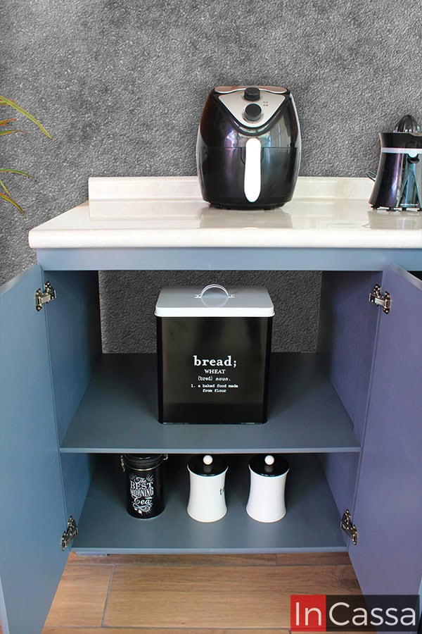 La foto muestra el mueble carnicero gris de la cocina, abierto para revelar su capacidad de almacenamiento. Este gabinete cuenta con dos puertas que se abren hacia fuera, ofreciendo acceso a dos niveles de almacenamiento. La cubierta es de formaica estilo ojo de pájaro, perfecta para colocar electrodomésticos como una freidora y un exprimidor de jugos. El diseño práctico y organizado maximiza el uso del espacio disponible, haciendo que la cocina sea funcional y estéticamente agradable.