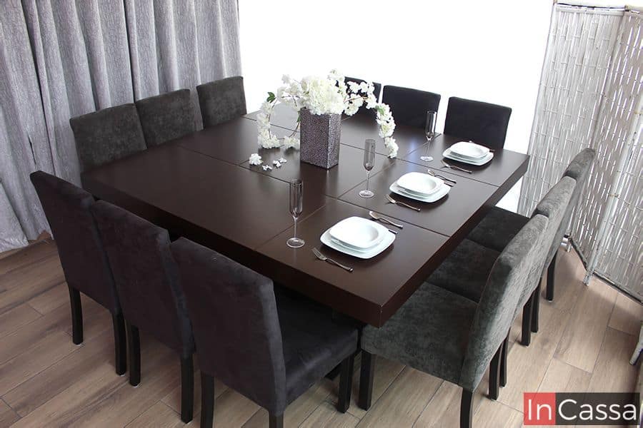 Una toma superior completa del comedor en su gran presentación para 12 personas. El conjunto incluye una amplia mesa en acabado tabaco mate y 12 sillas tapizadas en suede gris, todo presentado en una estancia decorada con una cortina gris y un biombo blanco.