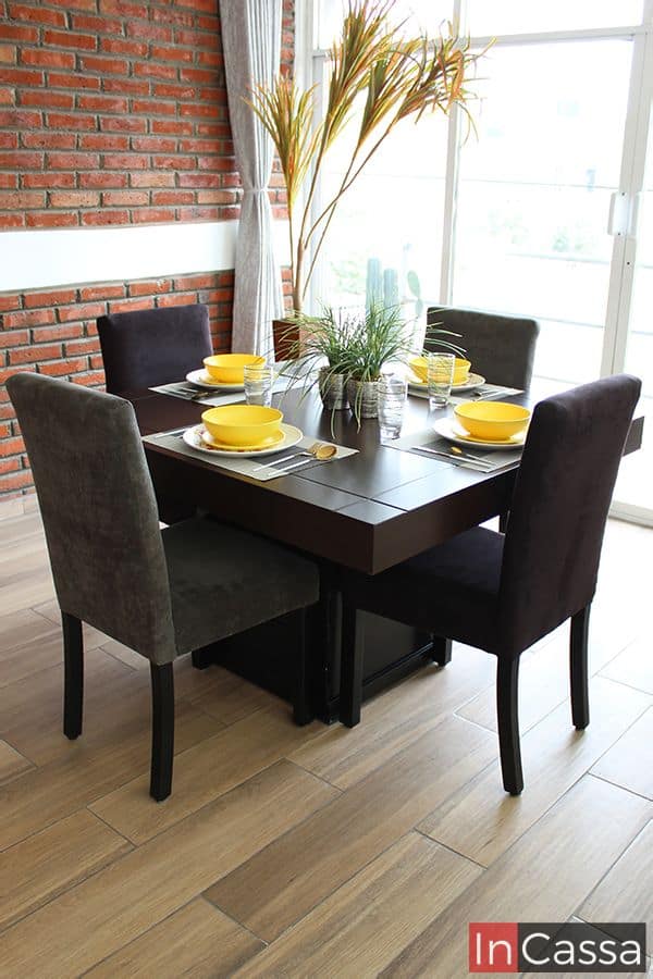Imagen del comedor en una práctica presentación de 4 sillas, cuya mesa es cuadrada con una cubierta en acabado tabaco y un pedestal de color chocolate, mientras que sus sillas están tapizadas en suede en tonos grises y chocolate. El conjunto se exhibe en una habitación con pared de ladrillo y piso de madera.