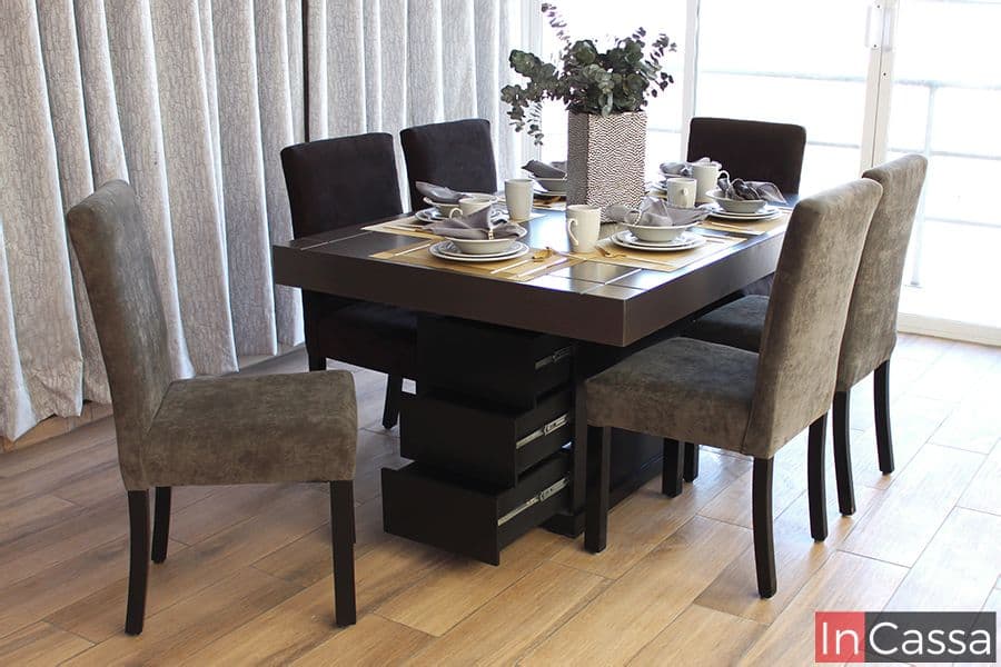 Fotografía del comedor presentado en su opción para 6 personas, en el que se destaca su pedestal chocolate con 3 cajones laterales integrados, al igual que sus sillas de suede en tonos grises y chocolate, mientras que la amplia cubierta de la mesa tiene un acabado tabaco mate.