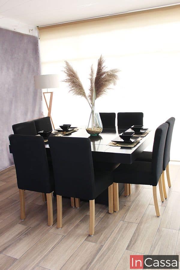 Foto del comedor moderno en su presentación para 8 personas. El conjunto se presenta en color negro completo exhibido en una estancia con pared gris, piso de madera y ventana completa.