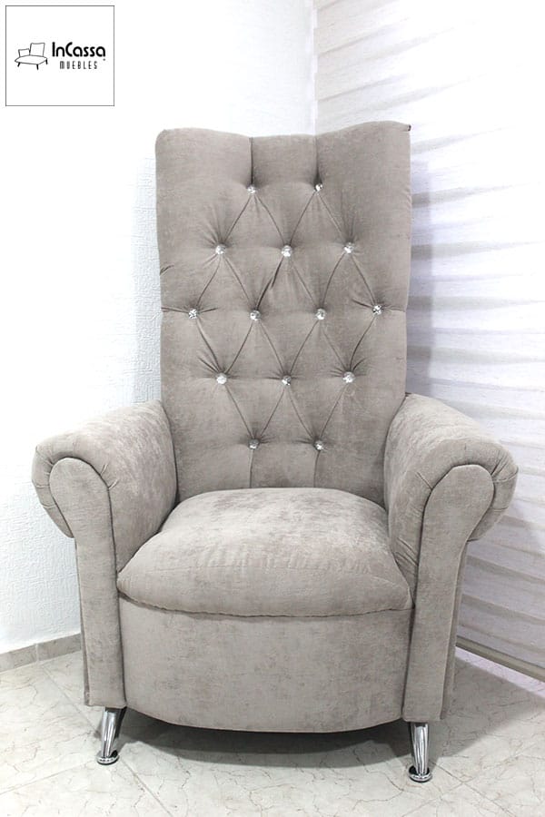 Este elegante sillón presenta un respaldo alto con capitoné y botones decorativos brillantes. Su tapizado en tela suede gris perla resalta su sofisticación, mientras que los brazos curvos y las patas cromadas añaden un toque moderno y refinado.