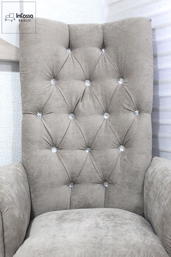 El respaldo alto de este sillón luce un diseño capitoné con botones decorativos en acabado cristal, realzando su elegancia. La tapicería de suede gris perla aporta suavidad y confort, haciendo de este mueble una opción sofisticada para cualquier espacio.