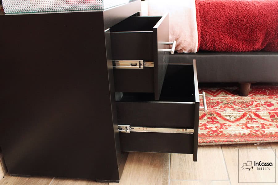 Se presenta un buró moderno con dos cajones abiertos que muestran correderas metálicas de extensión. El acabado chocolate resalta sobre el piso laminado y la alfombra roja, mientras que al fondo se distingue parte de la cama tapizada y la colcha en tono vino.