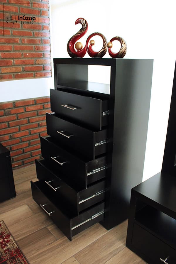 La imagen muestra una cajonera alta con sus cinco cajones abiertos y correderas metálicas de extensión, junto a un espacio abierto superior para exhibición. El acabado chocolate mate contrasta con la pared de ladrillo y el piso laminado, mientras figuras decorativas en tonos vino y dorado adornan la parte superior.