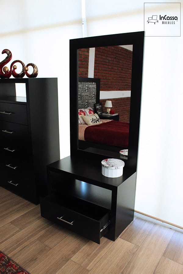 Se presenta un tocador moderno con gran espejo rectangular enmarcado en acabado chocolate mate. La base cuenta con una repisa intermedia y un cajón inferior con jaladera metálica. Sobre la superficie destaca una caja decorativa redonda, mientras el reflejo del espejo muestra parte de la cama y la pared de ladrillo.