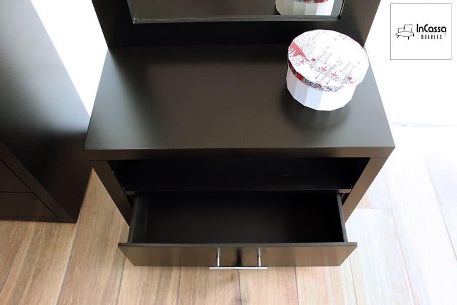 La imagen muestra una toma cenital del tocador en acabado chocolate mate, resaltando su repisa intermedia y cajón inferior con jaladera metálica. Sobre la superficie se aprecia una caja decorativa redonda con tapa estampada, mientras la luz natural realza el contraste entre el mueble y el piso de madera clara.