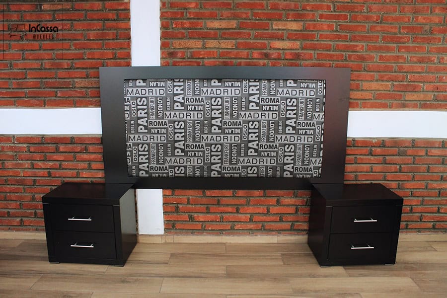 La foto presenta una toma frontal de la cabecera en acabado chocolate con panel central tapizado en lino ciudades, acompañada de dos burós laterales con doble cajón y jaladeras metálicas. El fondo de muro de ladrillo expuesto realza el contraste y estilo moderno del conjunto