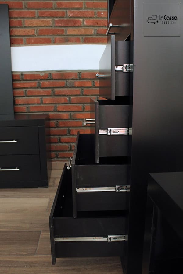 La imagen muestra una toma lateral de la cajonera en acabado chocolate con cuatro cajones abiertos, cada uno equipado con correderas metálicas de extensión y jaladeras rectas cromadas. Al fondo se aprecia parte de un buró y un muro de ladrillo, resaltando el estilo moderno del conjunto.