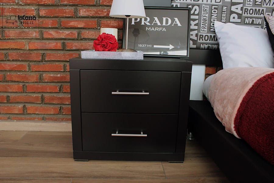 La foto presenta un buró moderno en color chocolate con dos cajones y tiradores metálicos rectos, ubicado junto a la cama. Sobre la superficie se aprecia una lámpara blanca, una caja decorativa con rosa roja y un cuadro enmarcado, todo frente a una pared de ladrillo aparente y sobre piso laminado.