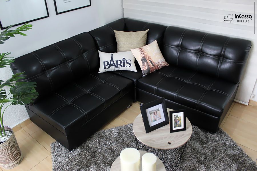 En esta foto se presenta la Sala Esquinera Modelo Londres, tapizada en tacto piel negro con costuras decorativas blancas en cuadros. Su diseño moderno y elegante se complementa con cojines decorativos, alfombra gris y mesas redondas, creando un ambiente acogedor y sofisticado.