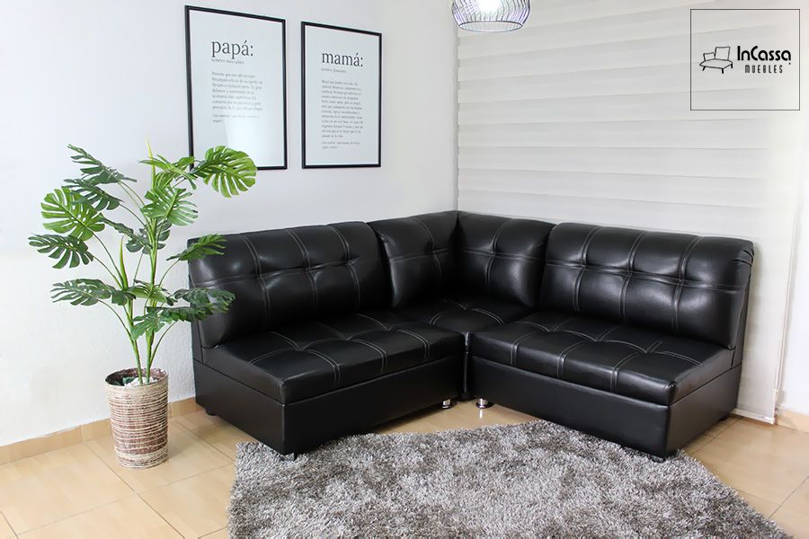 La foto presenta la Sala Esquinera Modelo Londres tapizada en tacto piel negro con costuras en diseño capitonado. Su forma en “L” aprovecha al máximo los espacios, ofreciendo asientos amplios y cómodos. La decoración minimalista con alfombra gris y planta natural realza el estilo moderno y elegante del conjunto.