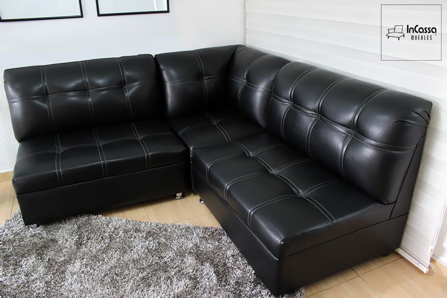 Se presenta la Sala Esquinera Modelo Londres en un ángulo lateral, mostrando su tapizado en tacto piel negro con costuras blancas en patrón cuadrado. El diseño en “L” brinda asientos amplios y confortables, mientras que la alfombra gris de textura suave complementa un estilo contemporáneo y elegante para cualquier sala moderna.