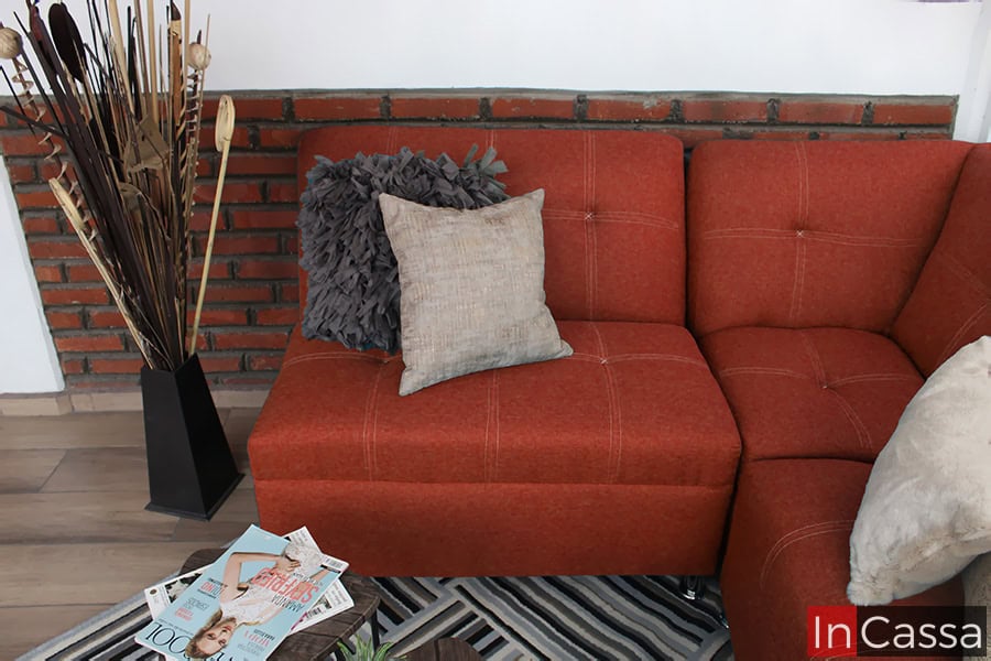 La foto presenta un acercamiento de la Sala Esquinera Modelo Londres tapizada en pliana color cobre. Se destacan las costuras en cuadros que realzan su diseño moderno, acompañadas por cojines decorativos en tonos gris y beige. La base metálica de las patas y la alfombra a rayas complementan un estilo cálido y sofisticado.