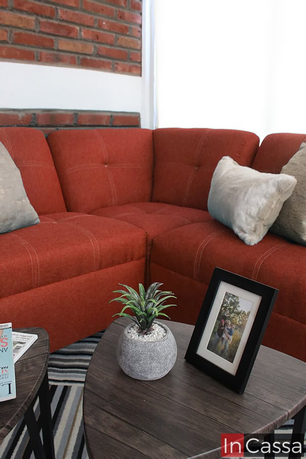 La imagen muestra un acercamiento a la esquina de la Sala Esquinera Modelo Londres, tapizada en pliana color cobre. Se aprecian las costuras decorativas en patrón cuadrado que aportan elegancia y modernidad, acompañadas por cojines en tonos claros y mesas auxiliares de madera con decoración minimalista.