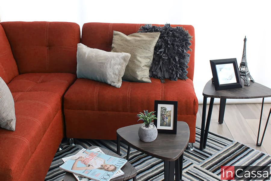 Se presenta una vista lateral de la Sala Esquinera Modelo Londres, tapizada en pliana color cobre. El diseño moderno resalta con costuras en cuadros y cojines decorativos en tonos gris y beige. Las mesas auxiliares triangulares en madera y la alfombra a rayas aportan un estilo contemporáneo y armonioso al espacio.