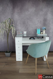 Mesa para uñas en color blanco con cuatro cajones en su pata izquierda (viéndola desde la parte de trasera); la mesa cuenta con una cubierta de cristal y un espacio para instalar una lampara de uñas. El mueble es exhibido en una habitación con pared gris y piso de madera.