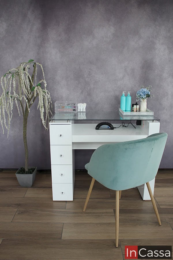 Mesa para uñas en color blanco con cuatro cajones en su pata izquierda (viéndola desde la parte de trasera); la mesa cuenta con una cubierta de cristal y un espacio para instalar una lampara de uñas. El mueble es exhibido en una habitación con pared gris y piso de madera.